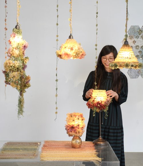 artist standing in room with hanging illuminated floral lampshades touching one of them and smiling