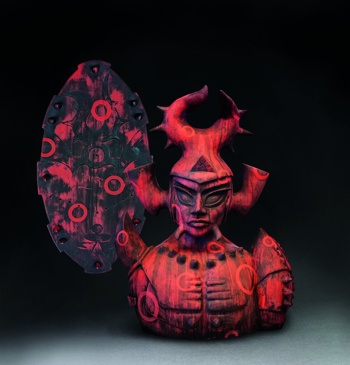 High fire clay and glazes creates a red clay piece resembling a warrior with a shield.