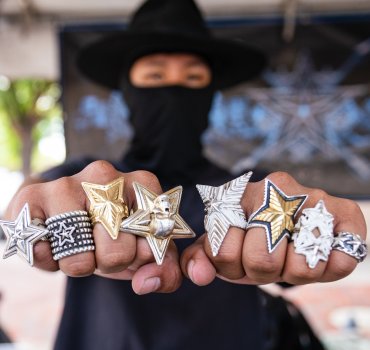 close-up of two fists adorned with metal star rings with person in the background wearing a black balaclava and wide-brimmed hat