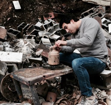 Ceramic artists seated among the rubble of a burned-down studio working on a clay jug on a broken wheel