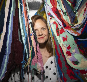 textile artist in studio peaking out from behind a pair of colorful tufted wall hangings