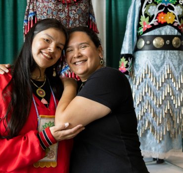 two women posing together with traditional jingle dresses on display behind them