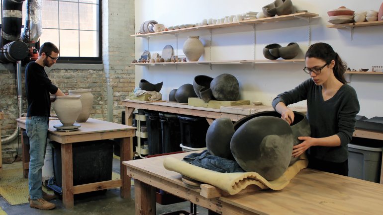 Two artists working on large ceramic vessels at respective tables in studio space with shelves