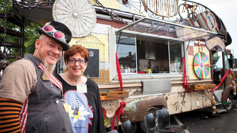 Two people one wearing circus inspired clothing posing in front of a circus-decorated food truck