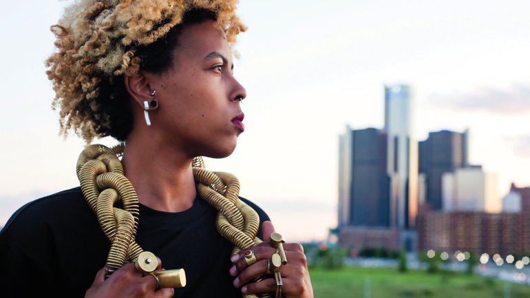 portrait of tiff massey with coiled gold necklace and large gold rings with detroit buildings in back on a summer evening