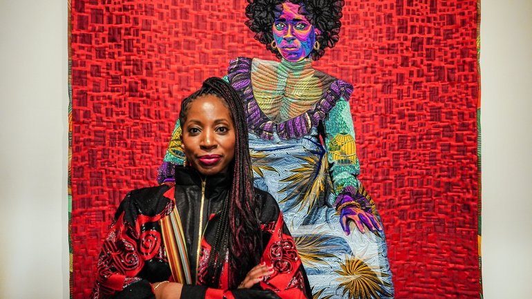 artist in red and black shirt posing in front of quilt depicting woman in vibrant colors over red backdrop