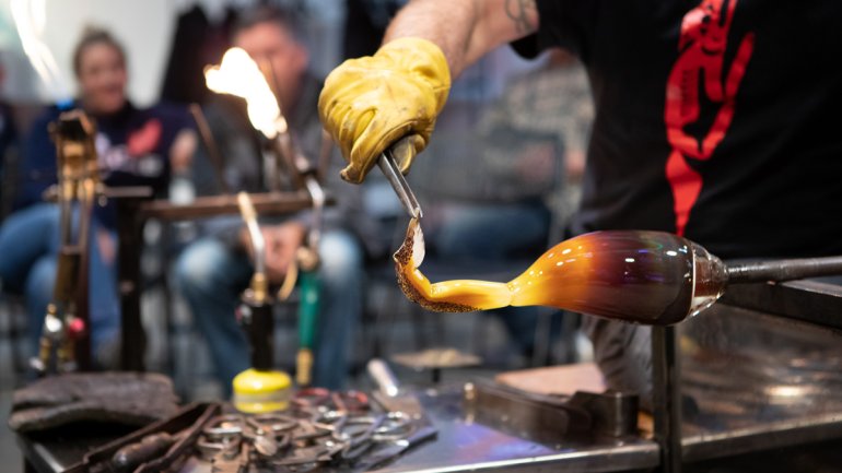 Person in black tshirt shaping hot glass as part of a demonstration in a workshop with onlookers