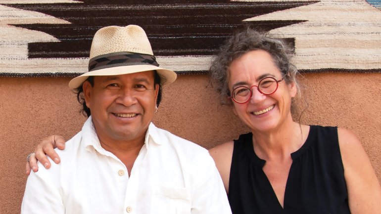 portrait of a man and women smiling and seated against and clay colored wall with black and white weaving hung on it