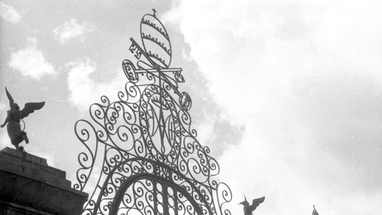 Black and white photograph of the ornate decorative crest of a wrought iron gate silhouetted against a cloudy sky