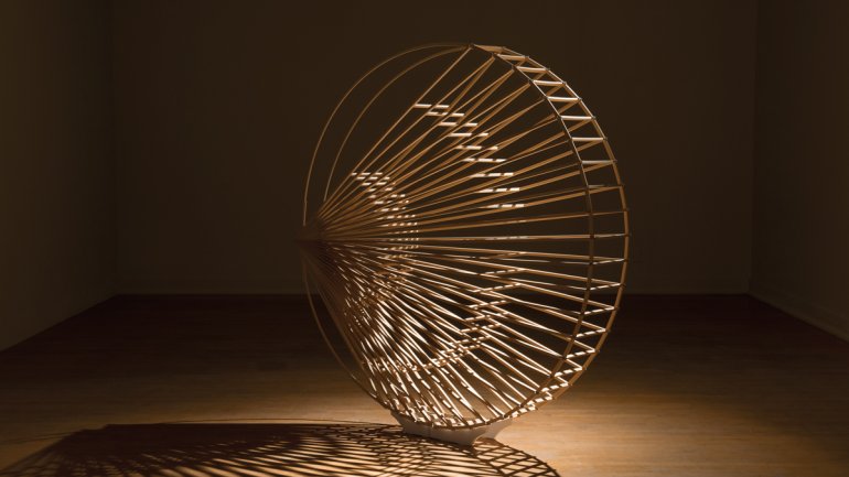Large coin-shaped wooden sculpture with inner spokes radiating from the left side pictured in an empty room with spotlighting