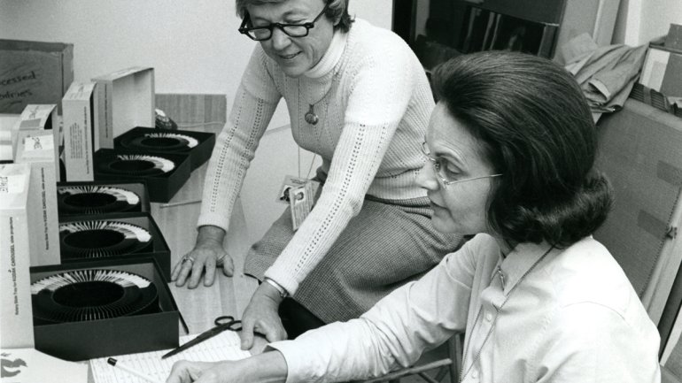 grayscale photo of two people in an office environment organizing rolls of slides