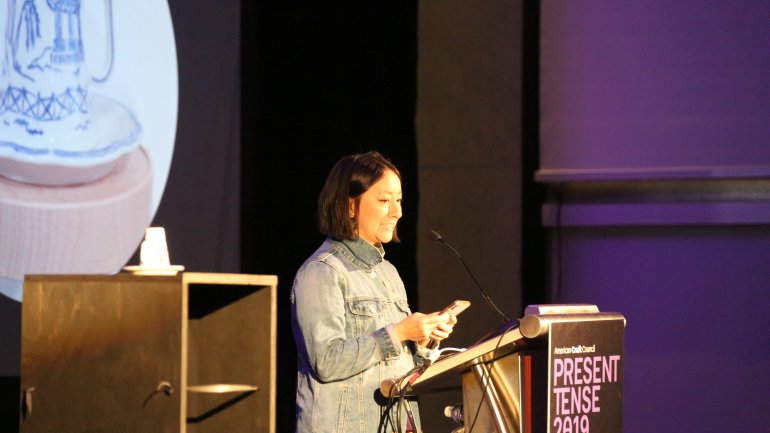 Person presenting at a podium beside a ceramic cup on a pedestal with slideshow image in backdrop