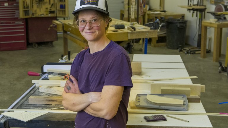 portrait of smiling woodworker leaning against shop table with crossed arms wearing hat and purple shirt