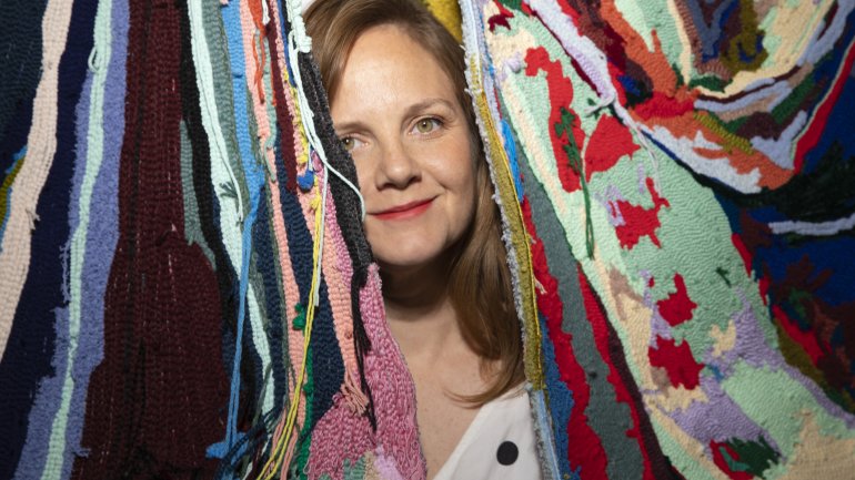 textile artist seated in studio beside colorful tufted wall hanging art