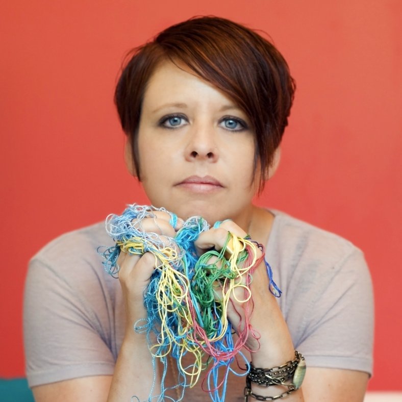 portrait of artist with chin on hands holding tangle of multi-colored embroidery thread with bright red background