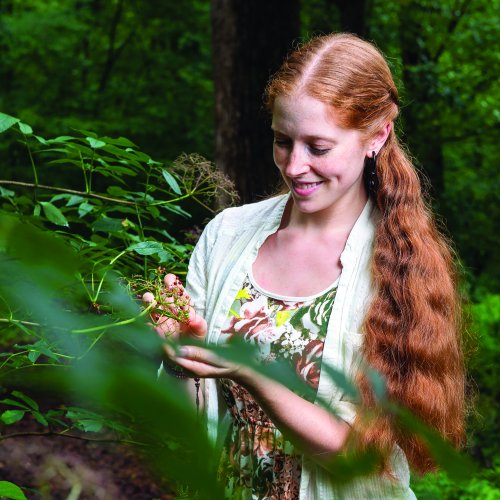 Portrait of person with long red hair examining flower in a forest