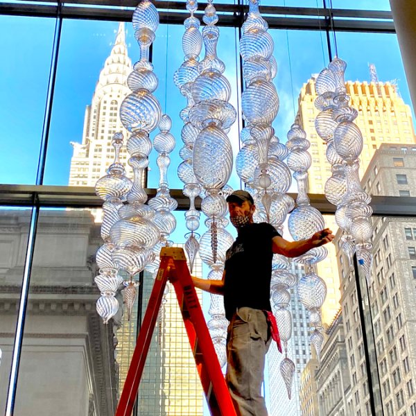 Glass artist posing on ladder while installing work in windowed gallery space in new york city