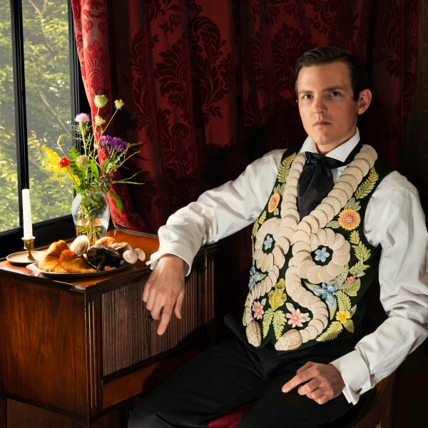 man in mid-19th-century garb posing in a chair wearing vest depicting flowers and snakes made from ceramic tiles