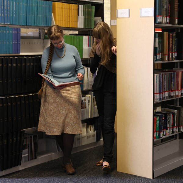 Two researchers looking at a book together in library stacks.