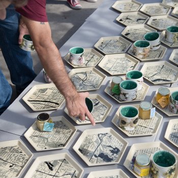 table with hexagonal ceramic plates with map illustrations topped with cup and honey jars with a person pointing at one of the plates