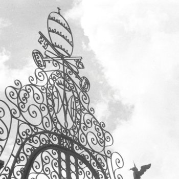 Black and white photograph of the ornate decorative crest of a wrought iron gate silhouetted against a cloudy sky