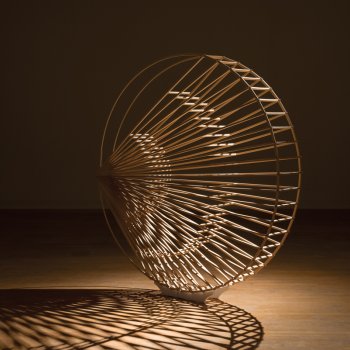 Large coin-shaped wooden sculpture with inner spokes radiating from the left side pictured in an empty room with spotlighting