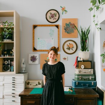 Portrait of Amanda McCavour posing against a desk in a studio with embroideries hanging on the walls