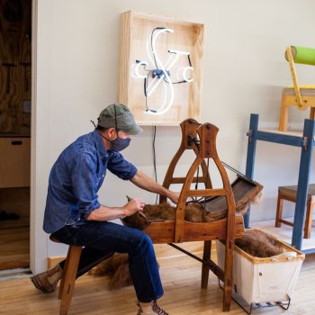 Upholstery artist seated working with an antique carding machine in a contemporary studio