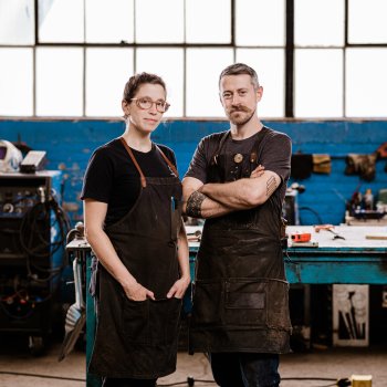 portrait of two blacksmiths wearing aprons posing in a shop with blue painted brick walls