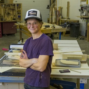 portrait of smiling woodworker leaning against shop table with crossed arms wearing hat and purple shirt