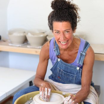ceramic artist in purple tank top and overalls working on a pottery wheel and smiling