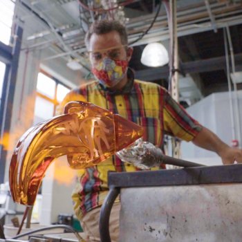 glass artists working an orange arrow-shaped glass sculpture with flame