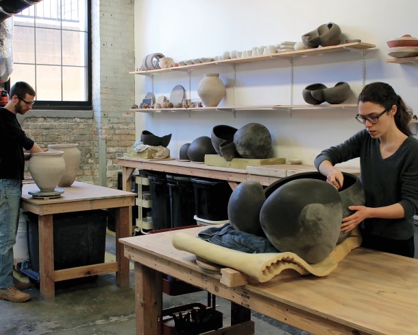 Two artists working on large ceramic vessels at respective tables in studio space with shelves