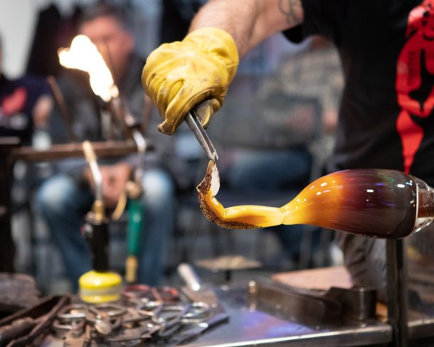 Person in black tshirt shaping hot glass as part of a demonstration in a workshop with onlookers