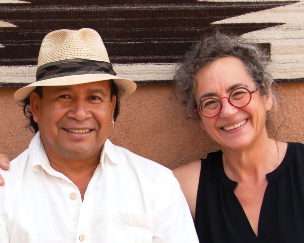 portrait of a man and women smiling and seated against and clay colored wall with black and white weaving hung on it