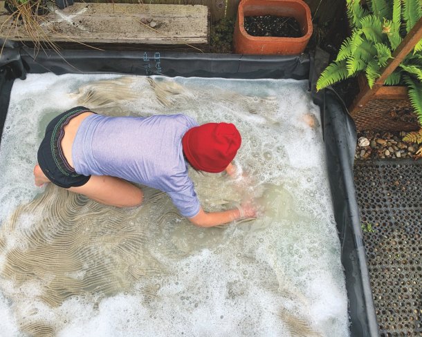 Artist kneeling a outdoor tub filled with soapy water massaging felt with hands