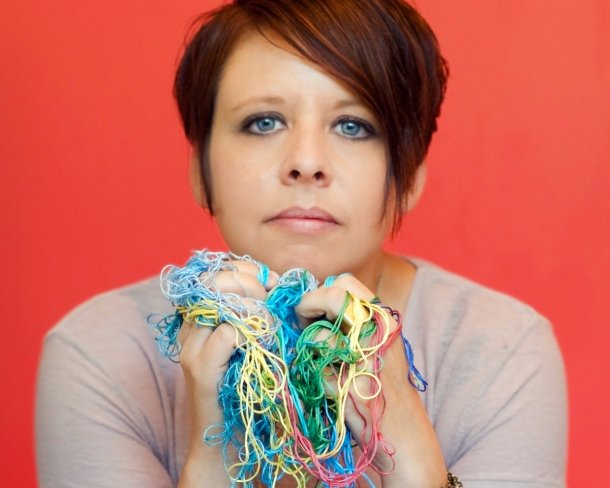 portrait of artist with chin on hands holding tangle of multi-colored embroidery thread with bright red background