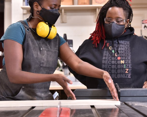 two women wearing face masks and protective glasses working in a shop at a table saw